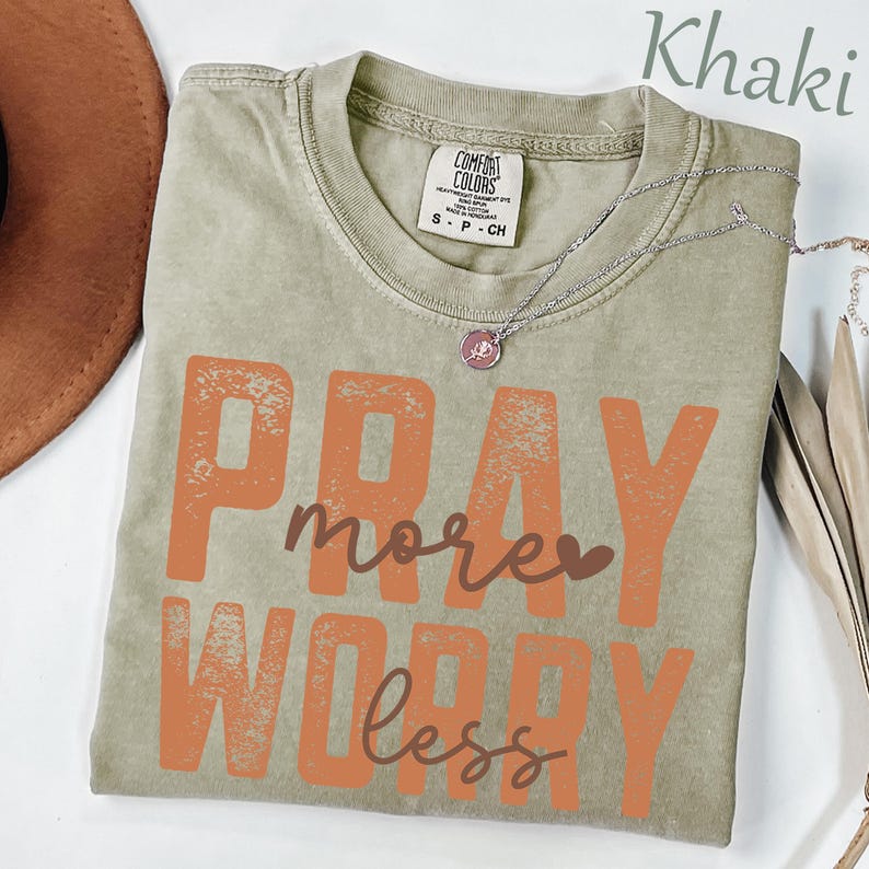 K&ouml;nnte beinhalten: Ein khakifarbenes Kurzarm-T-Shirt mit einem verwaschenen orangefarbenen Grafikdruck mit der Aufschrift PRAY more WORRY less. Das T-Shirt hat einen Rundhalsausschnitt mit einem Comfort Colors-Etikett und eine silberfarbene Kette.