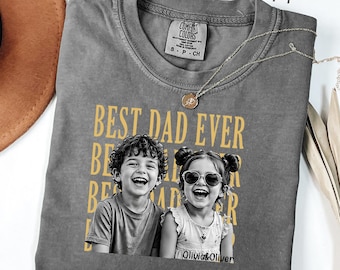 May include: A charcoal gray t-shirt featuring a yellow graphic with the text "BEST DAD EVER" repeated in the background. The center displays a black and white photograph of a young boy and a girl wearing heart-shaped sunglasses, with the text "Olivia&Oliver" below.