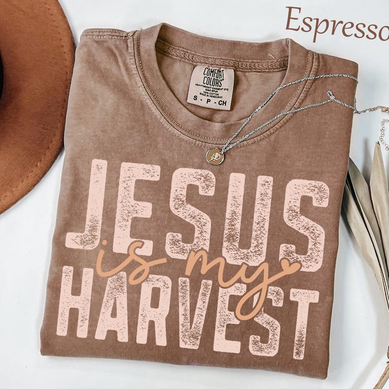 K&ouml;nnte beinhalten: Ein braunes Kurzarm-T-Shirt mit einem verwaschenen, hellrosa Grafikdruck mit der Aufschrift JESUS IS MY HARVEST. Das Shirt hat einen Rundhalsausschnitt und ein Comfort Colors Markenetikett am Innenkragen. Eine silberne Halskette liegt auf dem Stoff.