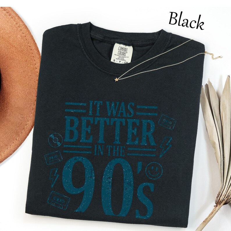 May include: A black crew neck t-shirt featuring the teal text IT WAS BETTER IN THE 90'S. The design includes graphic icons of a cassette tape, a lightning bolt, a smiley face, and a compact disc. The shirt has a Comfort Colors brand label.