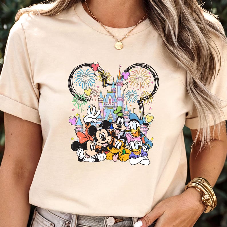 May include: Beige t-shirt featuring a colorful graphic of Disney characters, including Mickey and Minnie Mouse, Donald Duck, and Goofy. The design includes a castle, fireworks, and the iconic Mickey Mouse ears. The shirt is made of a soft material.