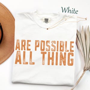 K&ouml;nnte beinhalten: Ein wei&szlig;es kurz&auml;rmeliges T-Shirt mit Rundhalsausschnitt und einem Comfort Colors Markenetikett am Innenkragen. Das Shirt zeigt den orangefarbenen Text ARE POSSIBLE ALL THING, der in einer abgenutzten Blockschrift auf der Brust aufgedruckt ist.