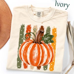 May include: An ivory t-shirt featuring a graphic of a large orange pumpkin with a brown stem and green leaves. The pumpkin is centered over vertical brushstroke stripes in shades of green, orange, and tan with white polka dots. The text Ivory is visible.