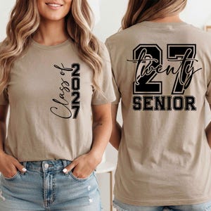 May include: Tan short-sleeved t-shirt with black text. The front of the shirt says "Class of 2027" vertically. The back of the shirt says "27 Twenty SENIOR" in a black and white design.