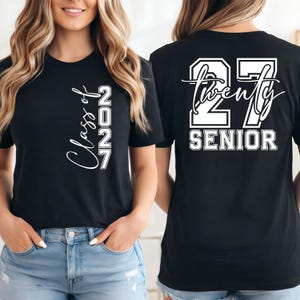 May include: Black t-shirt with white text. The front of the shirt says "Class of 2027" in a script font. The back of the shirt says "27 Twenty SENIOR" in a block font.