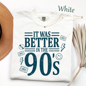 May include: A white short-sleeved t-shirt with a dark blue graphic print that reads IT WAS BETTER IN THE 90's. The design includes illustrations of cassette tapes, a lightning bolt, a smiley face, and a CD. The shirt features a Comfort Colors label.