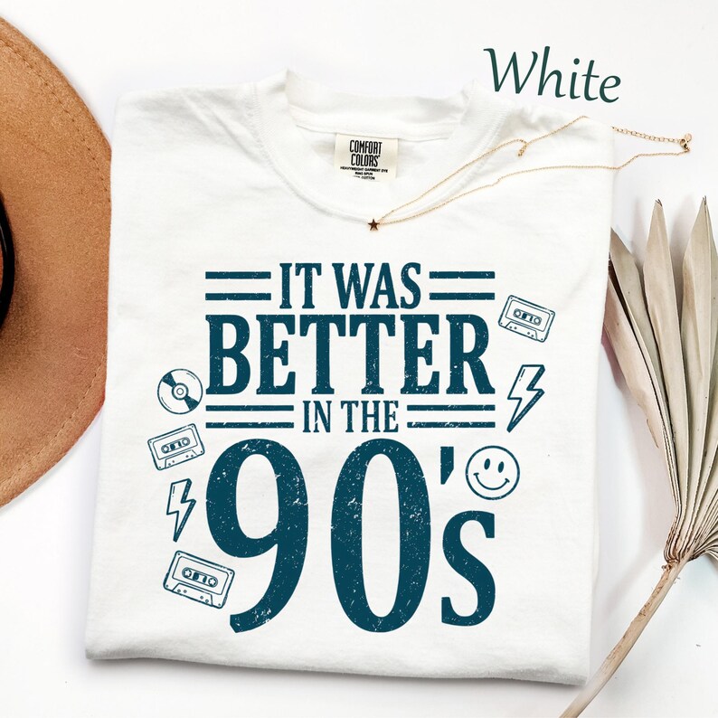 May include: A white short-sleeved t-shirt with a dark blue graphic print that reads IT WAS BETTER IN THE 90's. The design includes illustrations of cassette tapes, a lightning bolt, a smiley face, and a CD. The shirt features a Comfort Colors label.