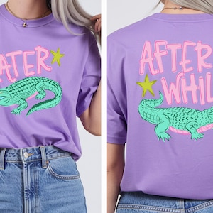 May include: A lavender t-shirt featuring a green alligator graphic and the text LATER on the front and AFTER WHILE on the back. The design includes pink and lime green star accents. The shirt has a crew neckline and short sleeves.