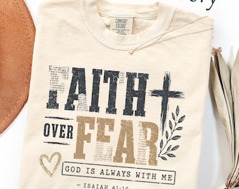 Peut inclure: Un t-shirt à manches courtes ivoire avec le texte FAITH OVER FEAR en lettres noires et dorées usées. Le motif comprend une croix, un cœur, des feuilles, ainsi que le texte GOD IS ALWAYS WITH ME et ISAIAH 41:10 avec des lignes décoratives.