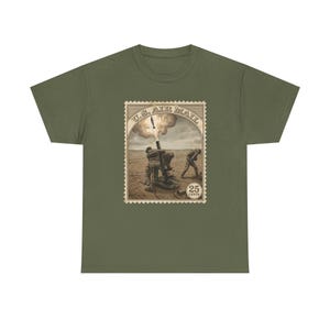 May include: Olive green t-shirt featuring a vintage U.S. Air Mail stamp design. The stamp depicts a sepia-toned scene of soldiers operating a cannon, with the text "U.S. AIR MAIL" at the top and "25 CENTS" at the bottom. The shirt is a classic crew neck.