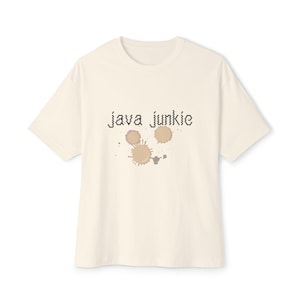 May include: Cream-colored t-shirt with the text "Java Junkie" in black pixelated font above three brown coffee stain graphics. The shirt has short sleeves and a crew neck.