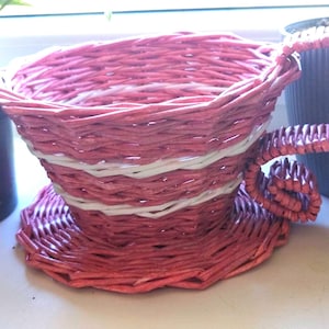 以下が含まれることがあります： 白いストライプのディテールが施された、織り込まれたピンク色のティーカップ型のバスケット。バスケットには、お揃いのソーサーベースと湾曲したハンドルが付いています。ティーカップは鉢植えの植物の隣にあります。