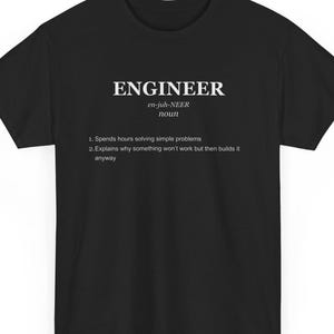 Può includere: T-shirt nera con la parola "ENGINEER" in bianco, la sua pronuncia e definizione. La definizione descrive un ingegnere come qualcuno che risolve problemi semplici e costruisce cose anche se sa che non funzioneranno.