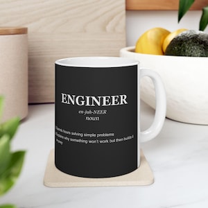 Op de afbeelding: Witte keramische mok met een zwarte buitenkant. De mok heeft het woord "ENGINEER" in het wit, samen met een definitie en humoristische uitspraken over ingenieurs. De mok staat op een beige onderzetter.