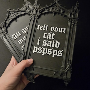 May include: Black gothic-style wall decor with white text that reads "tell your cat I said pspsps". The frame has ornate detailing. A hand holds the decor against a dark background.