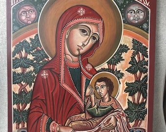 Puede incluir: Un icono religioso que representa una figura central con una túnica roja que sostiene a un niño. La obra de arte tiene un halo dorado y está sobre un fondo rojo oscuro. La pintura incluye texto y elementos decorativos.