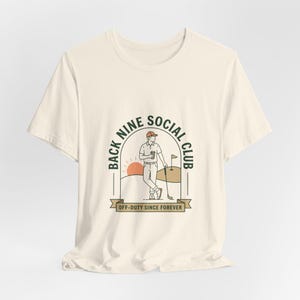 May include: Off-white t-shirt with a golf-themed illustration. The design shows a golfer, golf course details, and the text "BACK NINE SOCIAL CLUB" and "OFF-DUTY SINCE FOREVER". The graphic uses brown, orange, and green colours.