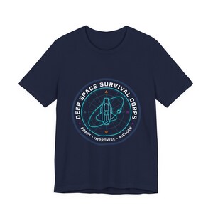 Peut inclure: T-shirt bleu marine avec un motif circulaire. Le motif présente un vaisseau spatial turquoise dans un cercle bleu et blanc, avec le texte "DEEP SPACE SURVIVAL CORPS". En dessous, les mots "ADAPT", "IMPROVISE" et "AIRLOCK".