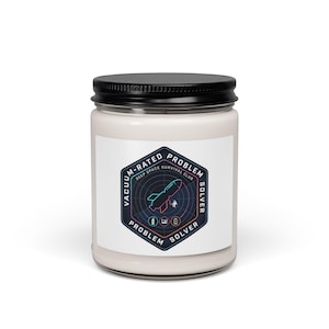 May include: A white candle in a glass jar with a black lid. The label features a hexagonal design with the text "Vacuum-Rated Problem Solver" and a rocket ship illustration. The candle is designed for deep space survival.