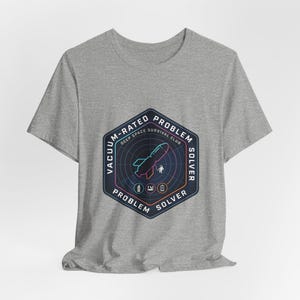 May include: A gray t-shirt featuring a hexagonal graphic with a rocket ship and the text "M-RATED PROBLEM SOLVER, DEEP SPACE SURVIVAL CLUB". The graphic has a blue, purple, and orange color scheme.