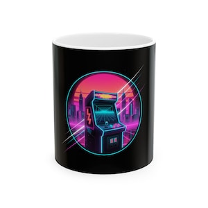 May include: Black and white ceramic mug featuring a retro arcade game design. The design includes a neon-lit arcade machine set against a cityscape with a pink and orange sunset. The mug has a white interior.