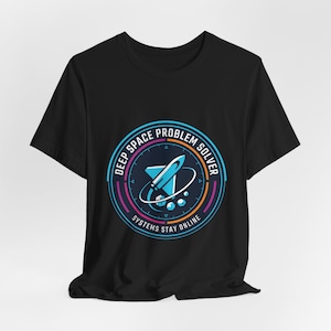 Pode incluir: Camiseta preta com um design gráfico circular que apresenta um foguete. O design inclui o texto "DEEP SPACE PROBLEM SOLVER" e "SYSTEMS STAY ONLINE" em tons de azul, rosa e laranja.