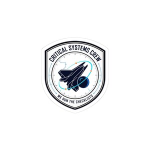 May include: A white sticker with a black and blue graphic of a space shuttle, a planet, and a curved line. The text "CRITICAL SYSTEMS CREW" and "WE RUN THE CHECKLISTS" are in a circular design.