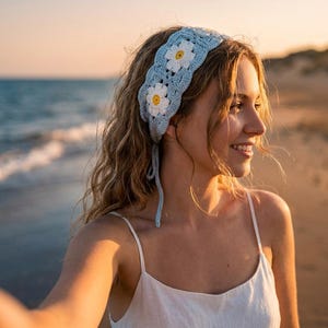 May include: A light blue crochet headband featuring a repeating pattern of white daisy motifs with yellow centers. The headband is constructed with intricate yarn stitching and includes thin, matching light blue ties that hang down from the sides.