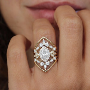 May include: A gold ring with a large oval-shaped diamond surrounded by smaller diamonds. The ring has a geometric design with a pointed top and a wide band.