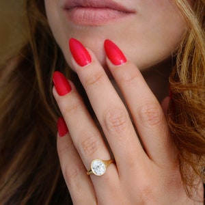 Op de afbeelding: Een gouden ring met een grote, ronde, heldere edelsteen. De ring zit aan een vinger met rode nagellak.