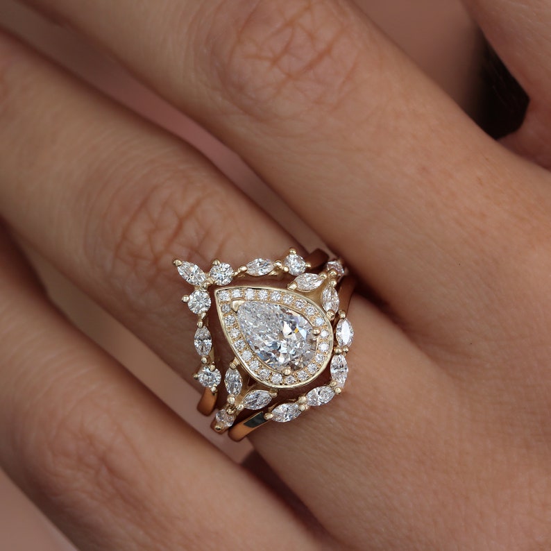Pear diamond three-ring set, the center stone is a 1.0-carat pear natural diamond or lab diamond. the engagement ring has a diamond halo and marquise diamonds on the band. The engagement ring has two nesting crown chevron wedding rings. solid gold