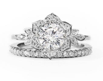 Puede incluir: Un anillo de compromiso de plata con un gran diamante redondo central rodeado de diamantes más pequeños en un diseño floral. La banda también está engastada con diamantes más pequeños.