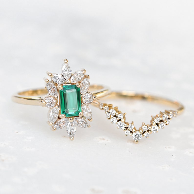 May include: A gold ring with a large emerald green gemstone surrounded by smaller diamonds. The ring is paired with a matching gold band with a diamond-encrusted design.