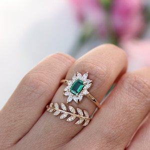 Puede incluir: Dos anillos de oro. Uno presenta una gema verde esmeralda rectangular rodeada de gemas blancas. El segundo anillo tiene un diseño de hojas con pequeñas gemas blancas. Los anillos están en un dedo.