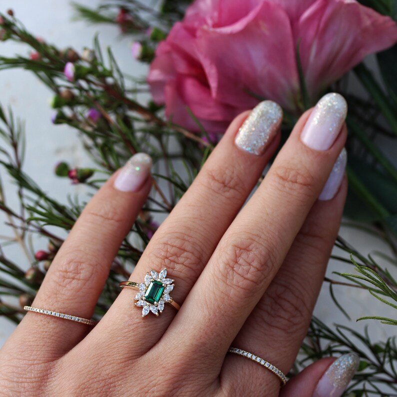 May include: A gold ring with a large emerald green gemstone surrounded by smaller diamonds. The ring is on a finger with sparkly nail polish.