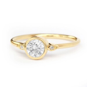 Könnte beinhalten: Ein goldener Ring mit einem runden, gefassten Diamanten und zwei kleineren Akzentdiamanten auf der Ringschiene.