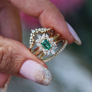 May include: A gold ring with a large emerald green gemstone surrounded by smaller diamonds. The ring is set on a band with a delicate diamond pattern.