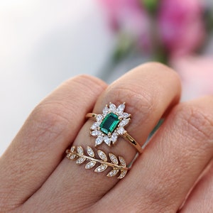 May include: A gold ring with a large emerald green gemstone surrounded by smaller clear gemstones. The ring is on a finger with a second gold ring with a leaf design.