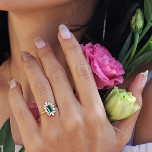 May include: A gold ring with a green emerald gemstone surrounded by smaller diamonds. The ring is on a woman's finger.
