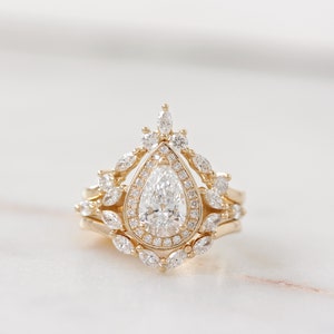 Pear diamond three-ring set, the center stone is a 1.0-carat pear natural diamond or lab diamond. the engagement ring has a diamond halo and marquise diamonds on the band. The engagement ring has two nesting crown chevron wedding rings. solid gold