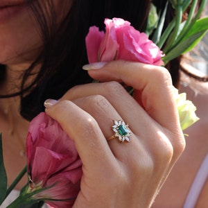 May include: A gold ring with a large emerald green gemstone surrounded by smaller diamonds. The ring is on a person's finger, and they are holding pink flowers.