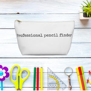 Op de afbeelding: Een wit etui met ritssluiting en de zwarte tekst Professional pencil finder in het midden gedrukt. Het etui heeft een goudkleurige metalen ritssluiting en een glad, schoon buitenoppervlak voor het opbergen van kleine kantoorartikelen.