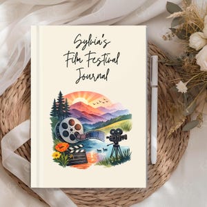 May include: A cream-colored journal with the text Sylvia's Film Festival Journal printed in black. The cover features a colorful illustration of a film reel, a movie camera, a clapperboard, and a landscape with mountains, trees, and a sunset.