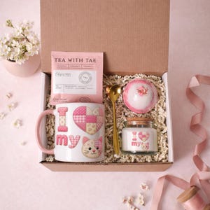 Puede incluir: Una caja de regalo con temática rosa, que contiene una taza con "I love my" y un diseño de gato, un sobre de té, una bomba de baño, una vela y una cuchara dorada. La taza tiene un asa rosa y la caja está llena de papel triturado.