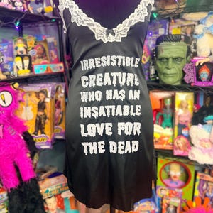 May include: A black sleeveless nightgown featuring a white lace trim along the V-neckline with a small black bow. The front displays the white text: IRRESISTIBLE CREATURE WHO HAS AN INSATIABLE LOVE FOR THE DEAD in a distressed, jagged font style.
