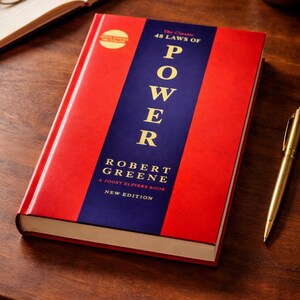 Puede incluir: Un libro de tapa dura titulado "The 48 Laws of Power" de Robert Greene. El libro tiene una cubierta roja con una franja vertical azul en el centro, que muestra la palabra "POWER" en letras blancas grandes. Tambi&eacute;n se ve un bol&iacute;grafo y gafas doradas.