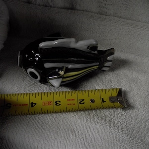 Puede incluir: Una figura de pez de cerámica en blanco y negro con detalles amarillos en las aletas laterales. El pez está colocado junto a una cinta métrica amarilla, que indica que el objeto mide aproximadamente 12,7 cm de largo total de cabeza a cola.