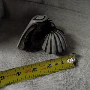 Puede incluir: Una figura de pelícano de cerámica con esmalte blanco y marrón oscuro y un cuerpo acanalado en forma de concha. Una cinta métrica amarilla está colocada frente a la figura, mostrando que mide aproximadamente 8,9 cm de longitud total.