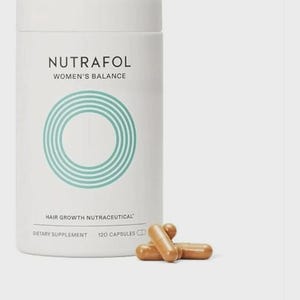 May include: A white cylindrical bottle of Nutrafol Women's Balance hair growth supplement with a teal circular design. The bottle is labeled "Hair Growth Nutraceutical" and "Dietary Supplement, 120 Capsules." Several brown capsules are next to the bottle.