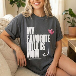 May include: Dark gray t-shirt with the text "MY FAVORITE TITLE IS MOM" in white block letters. Two pink hearts are above the word "MOM". The shirt has rolled-up sleeves.